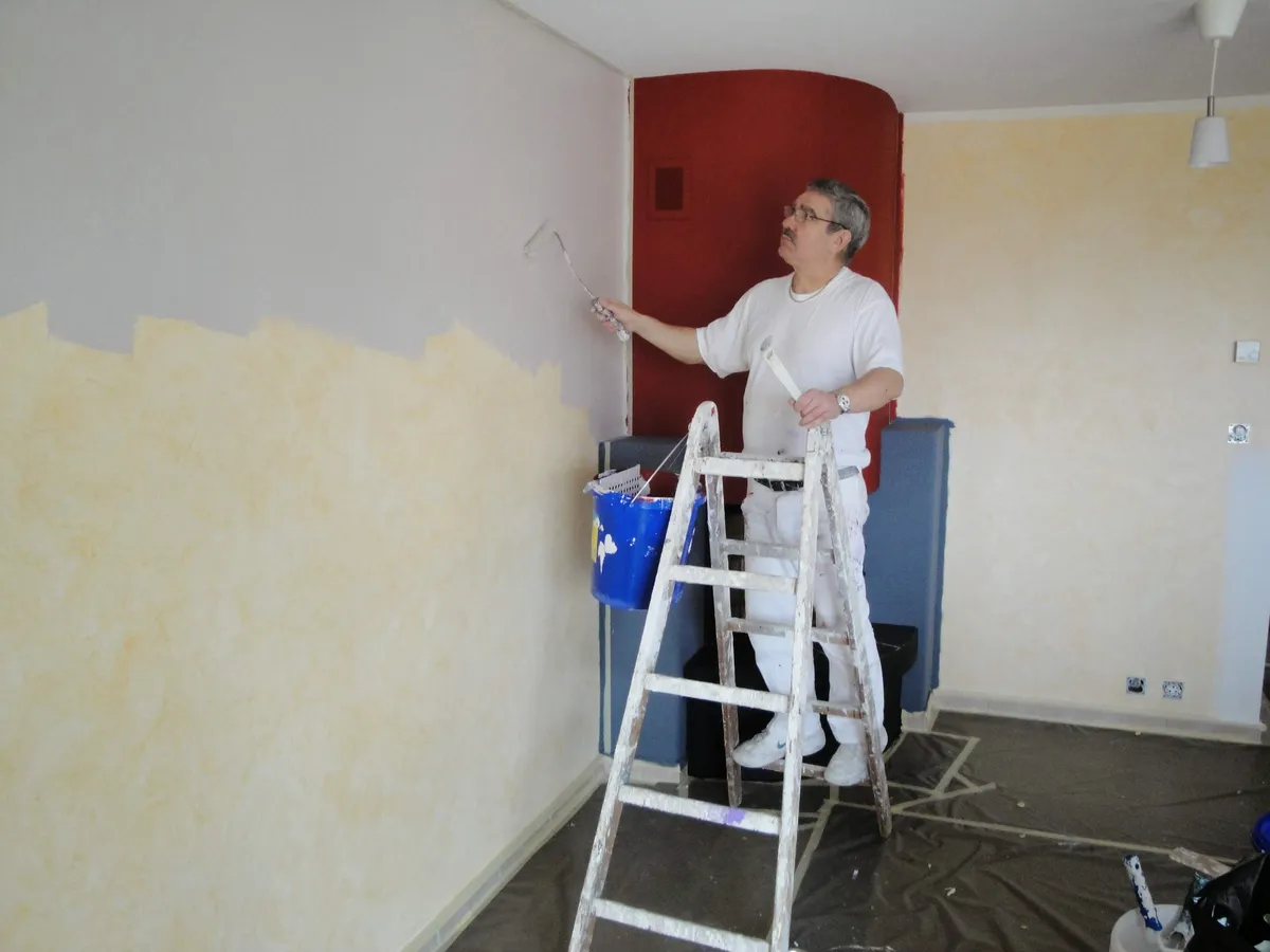 Application d'un enduit décoratif jaune à l'échelle, près d'un poêle rouge, une finition à effet matière signée l'atelier Peinture Casabene.