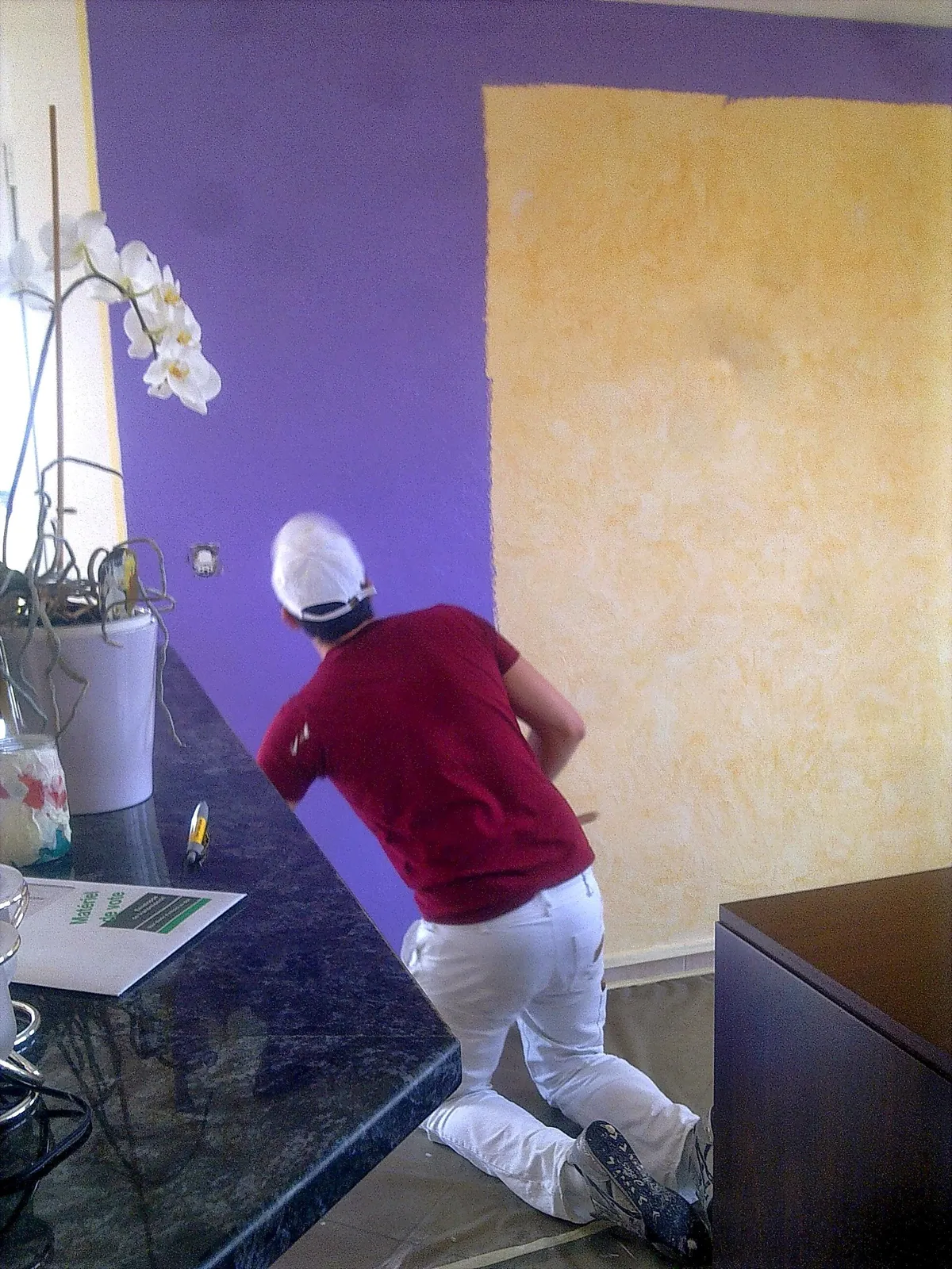 Peintre appliquant un enduit décoratif jaune mat sur un mur violet dans une cuisine ouverte.