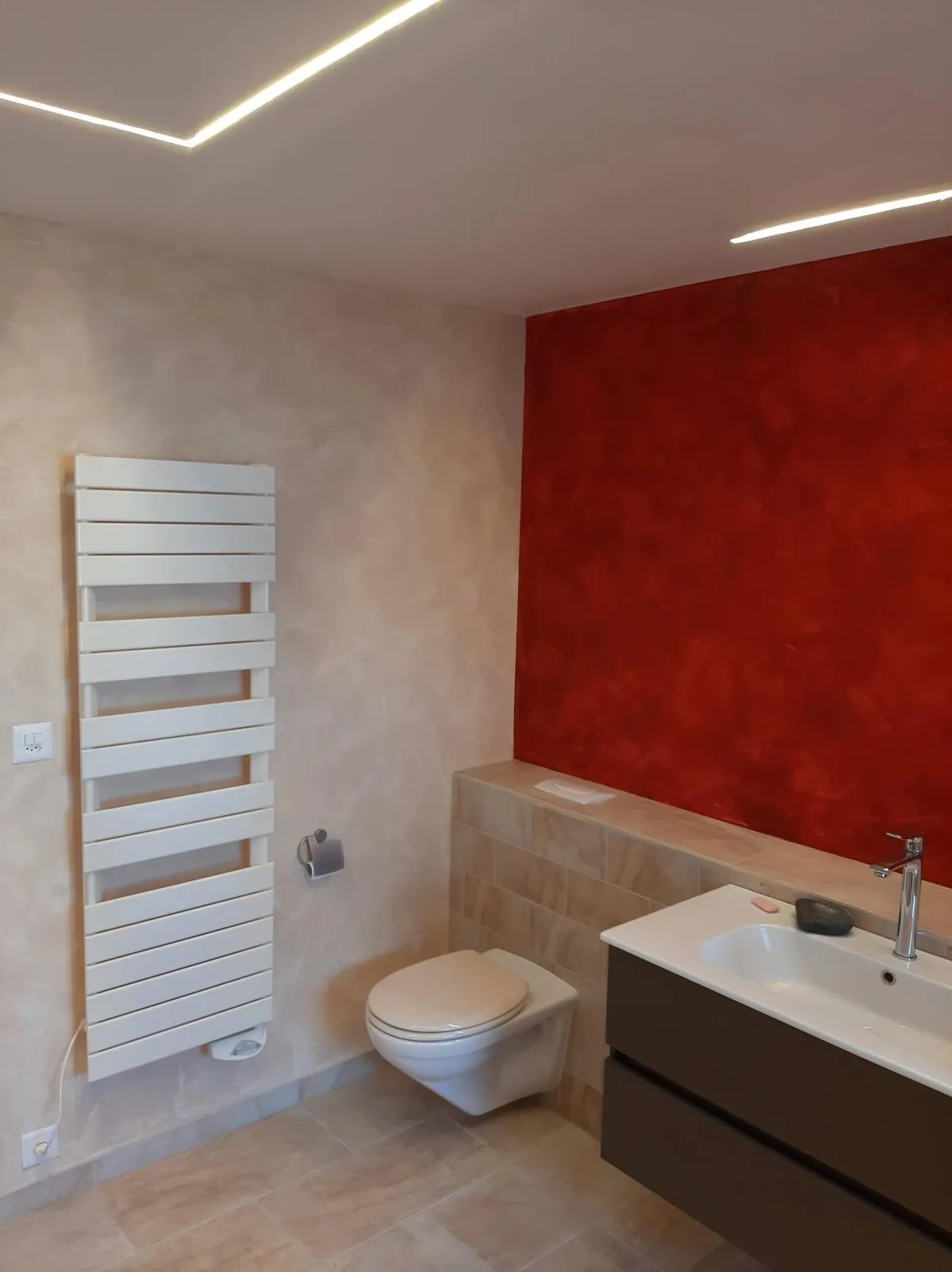 Salle de bain en enduit décoratif rouge et taupe, radiateur sèche-serviettes blanc et éclairage linéaire encastré au plafond.