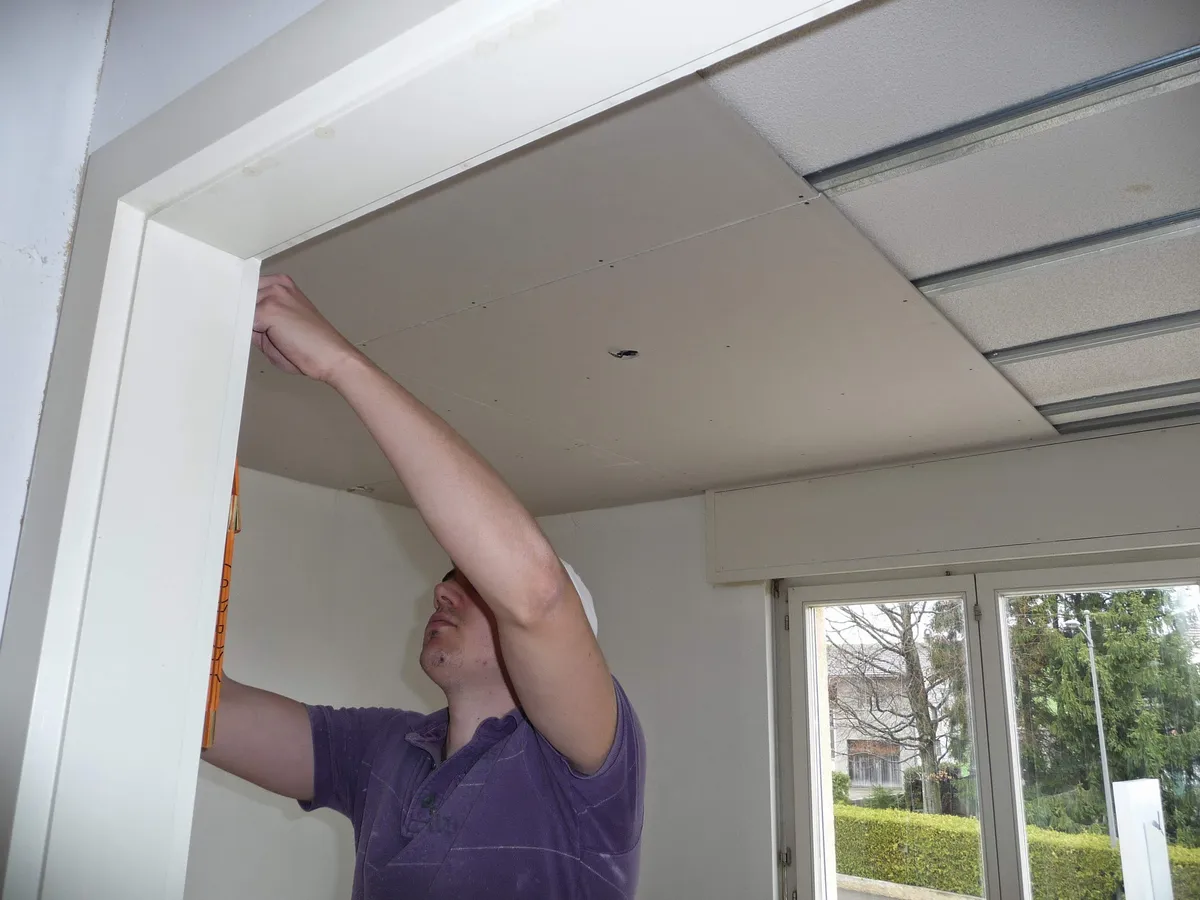 Fixation d'un panneau de faux plafond au-dessus d'une porte, avec le jardin visible en arrière-plan.