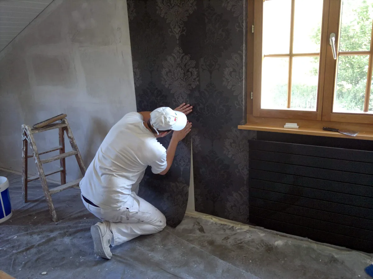 Pose de papier peint à motif damassé gris près d'une fenêtre, avec un radiateur noir installé sous l'appui.