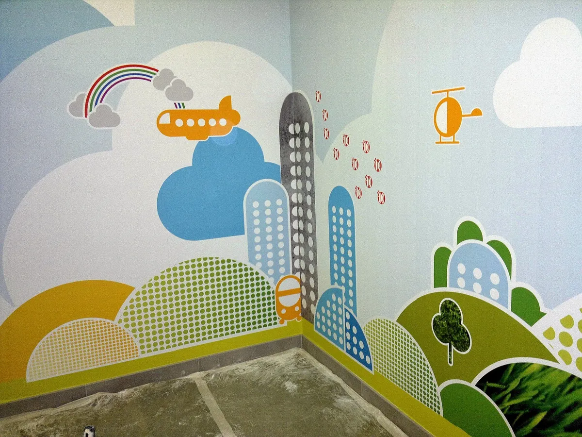 Coin de chambre d'enfant tapissé d'un papier peint panoramique représentant une ville fantaisiste et un arc-en-ciel.