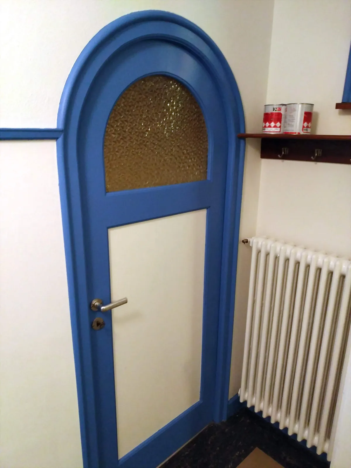 Porte arrondie bleue à vitrail dépoli surplombant un radiateur, une peinture de boiseries au-dessus d'un sol en marbre noir.