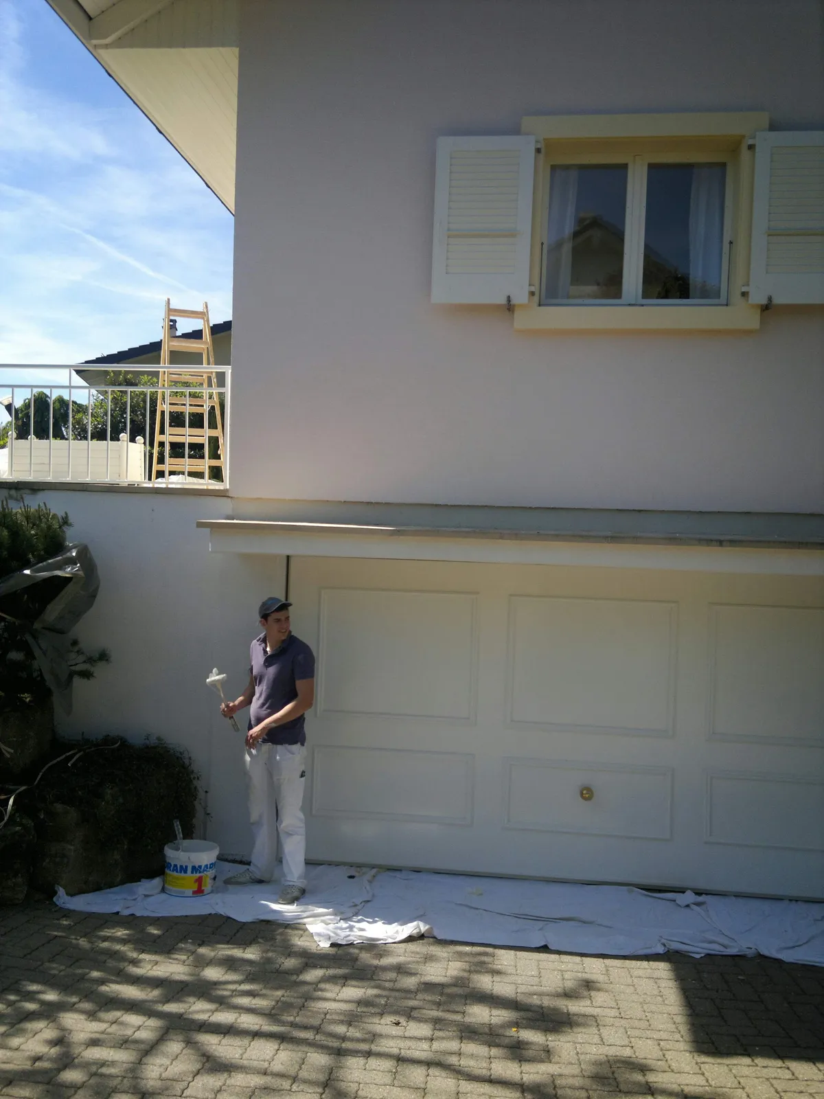 Peintre appliquant une peinture blanche sur une porte de garage, avec une échelle posée sur le balcon.