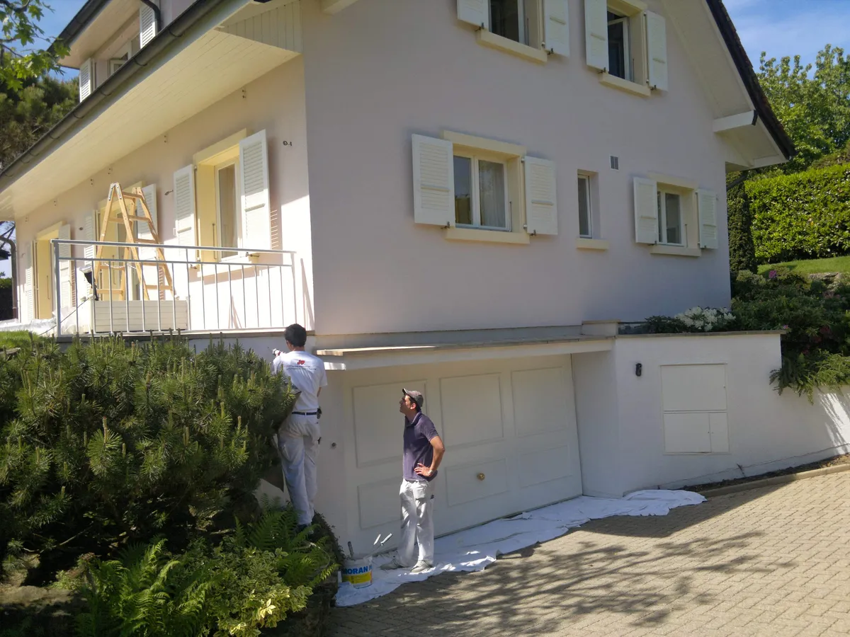 Peintre appliquant au rouleau la peinture d'une façade au-dessus d'un garage, buissons taillés au premier plan.