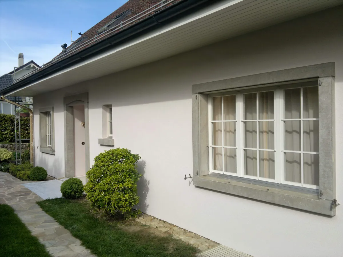 Façade beige fraîchement repeinte, avec des fenêtres à volets et un jardin fleuri.