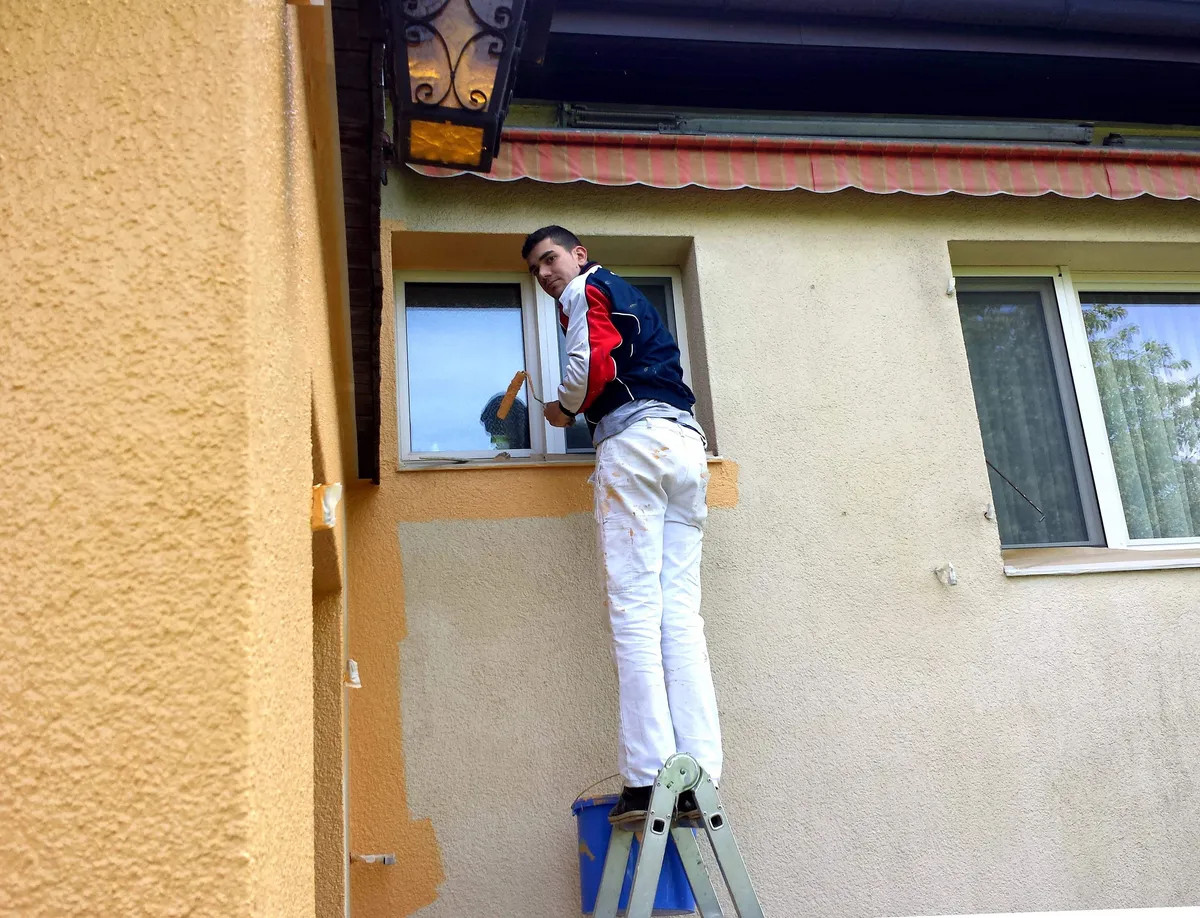Peintre sur échelle repeignant une façade bicolore jaune et beige près d'une fenêtre.