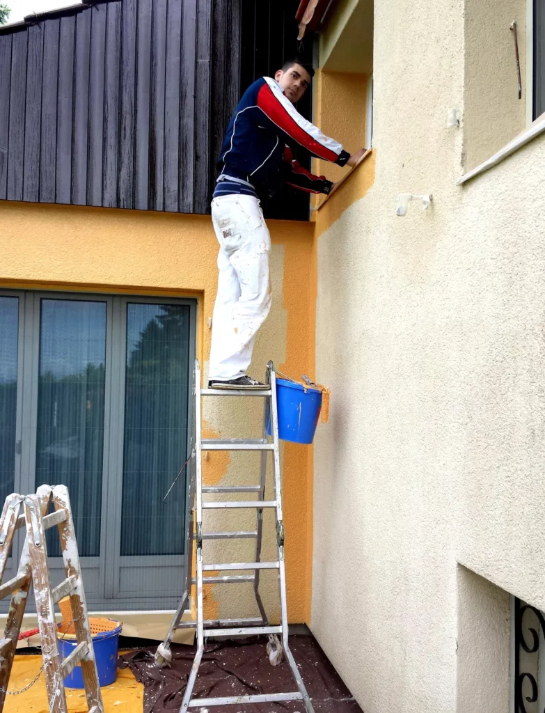 Peintre sur échelle repeignant l'angle d'une façade jaune, avec des seaux de peinture posés au sol.