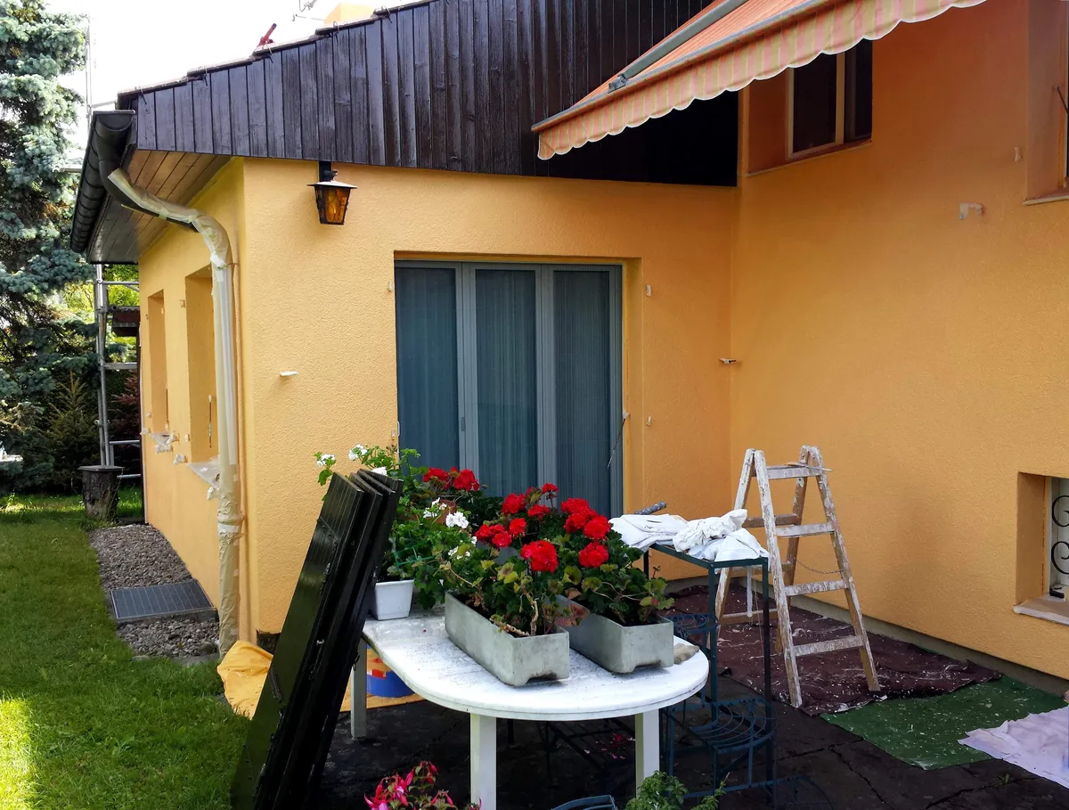 Terrasse avec façade jaune fraîchement repeinte, table fleurie et échelle rangée contre le mur, fin de ce chantier de peinture de façade.