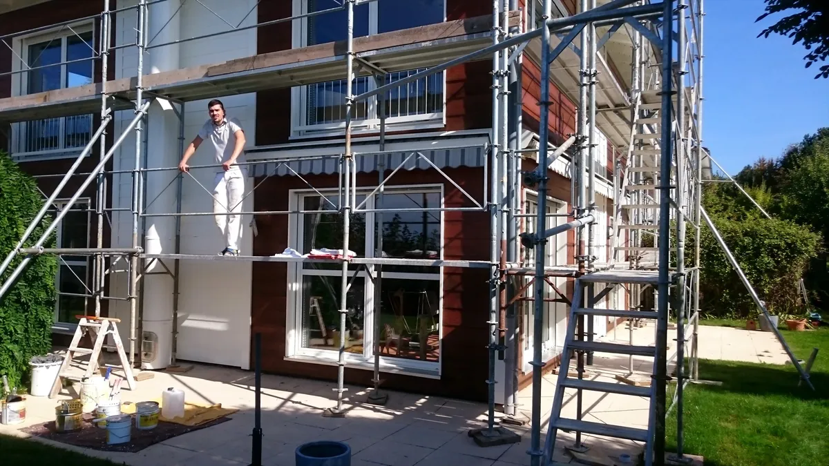 Peintre debout sur un échafaudage face à une façade rouge et blanche, avec un jardin en contrebas.