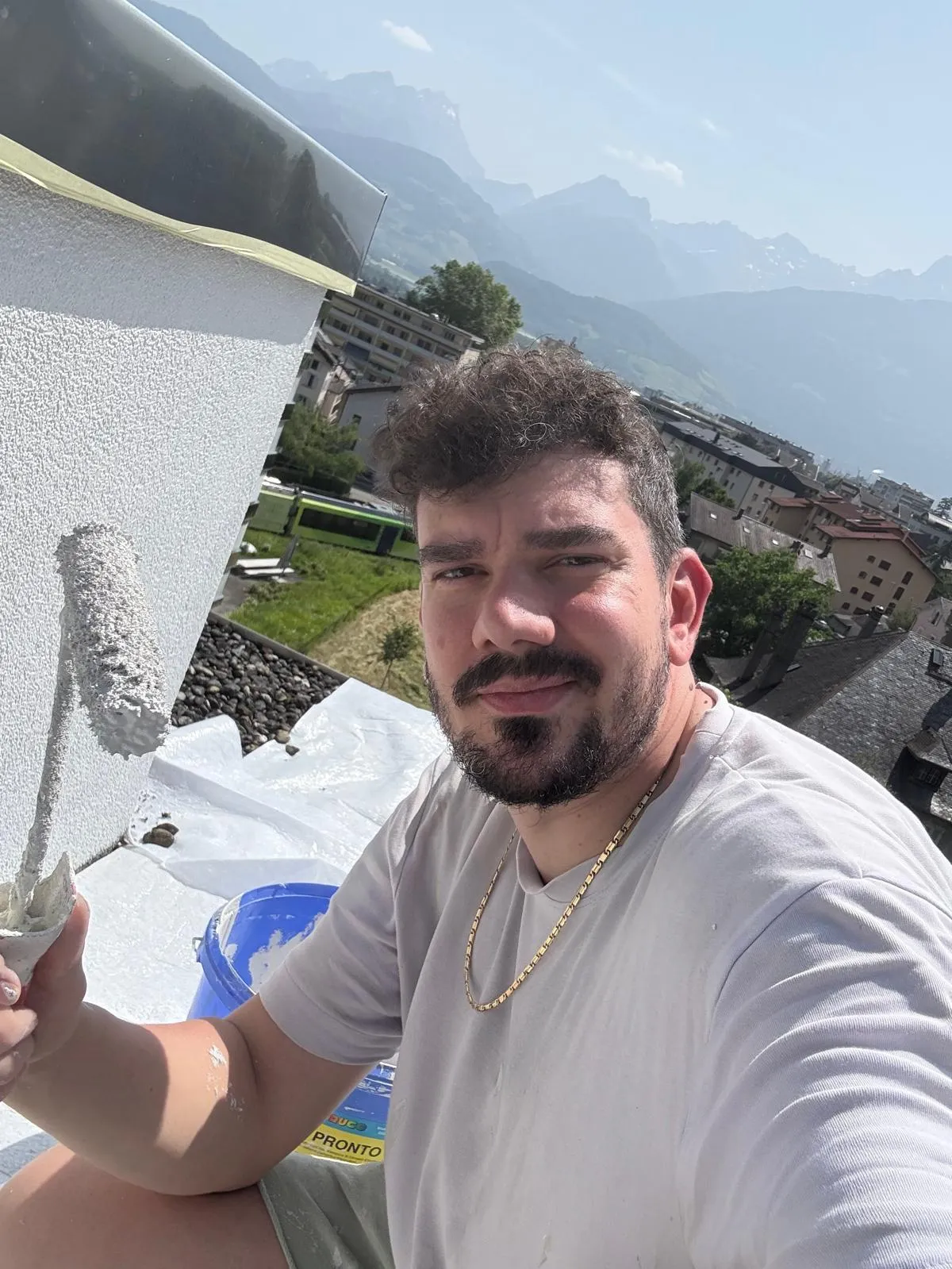 Paolo Casabene appliquant au rouleau une peinture de façade sur l'acrotère d'un attique, chantier en hauteur face aux montagnes.