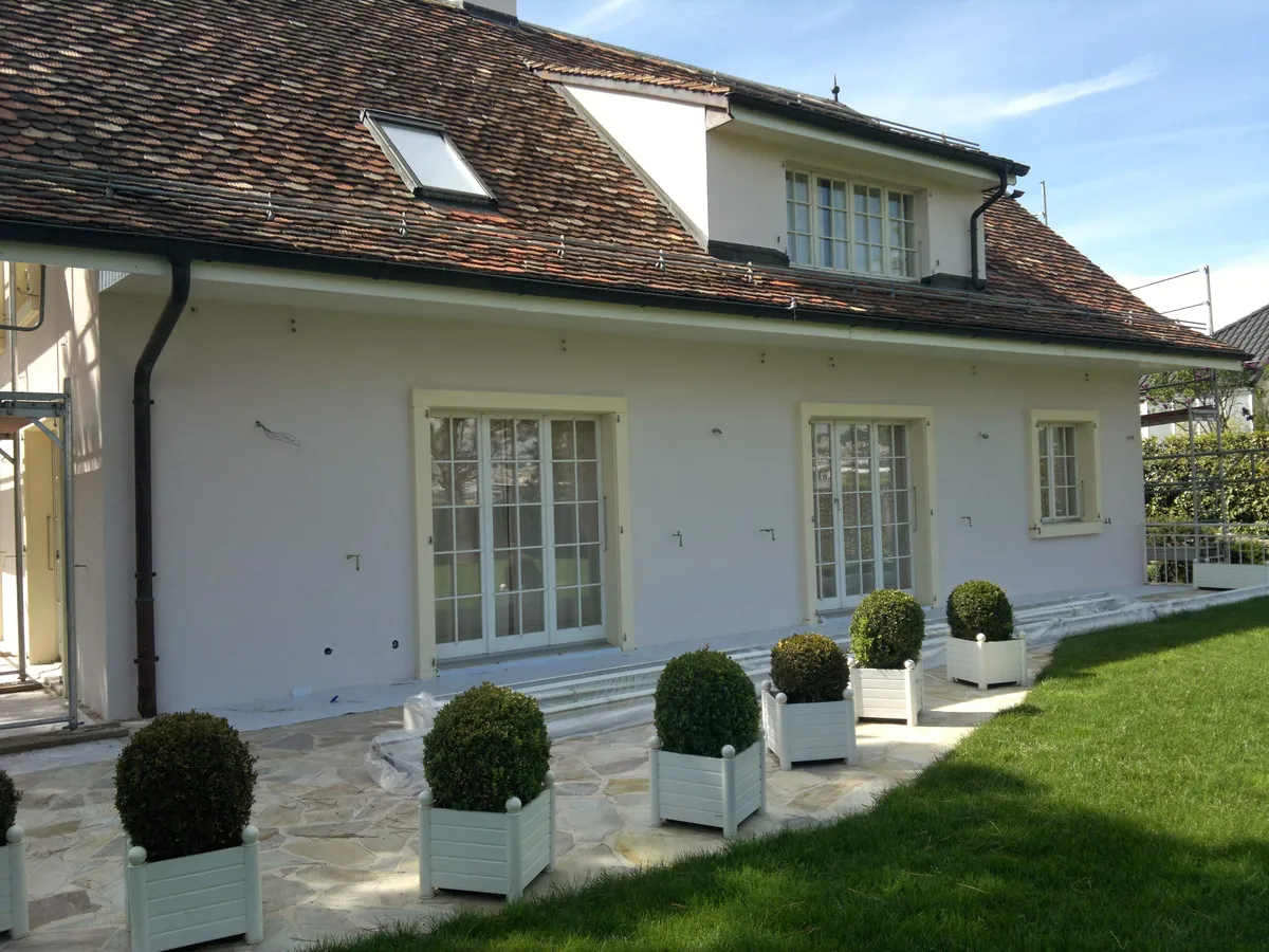 Façade repeinte en blanc cassé, terrasse dallée bordée de buis en pots, un chantier de peinture de façade terminé récemment.