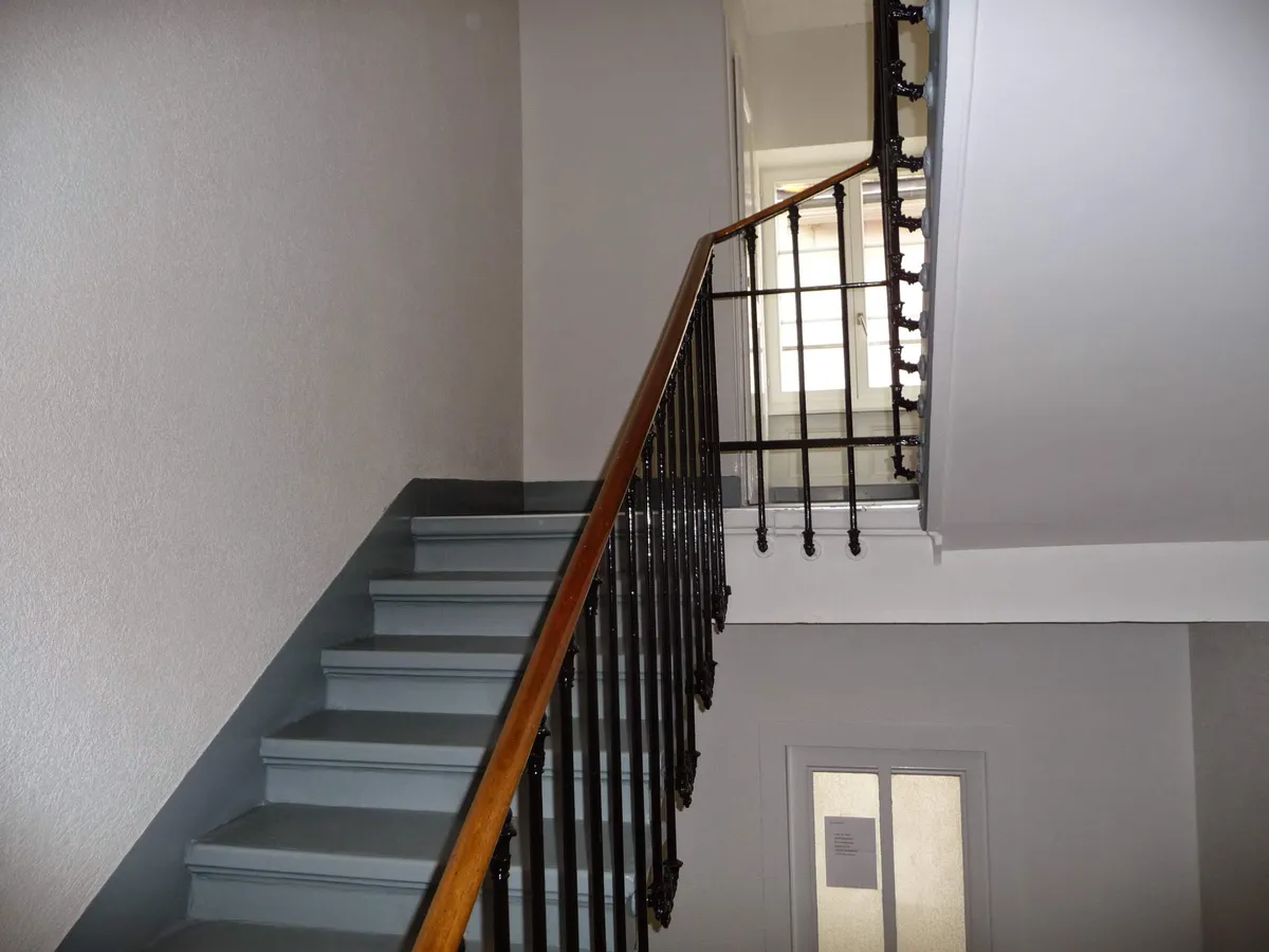 Cage d'escalier repeinte en blanc, avec une rampe en bois et un garde-corps en fer forgé.