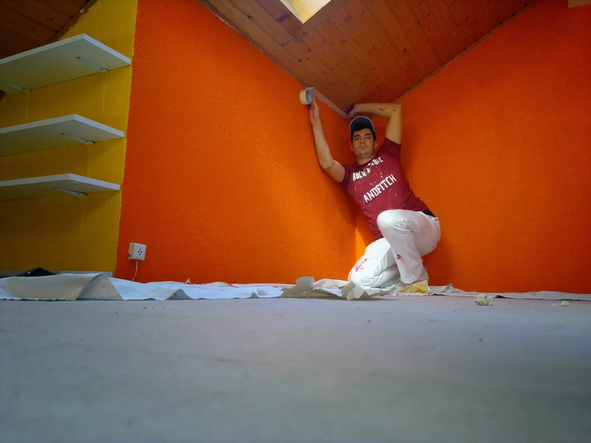 Peintre appliquant une peinture orange sous un plafond en pente, entre un mur jaune et des étagères.