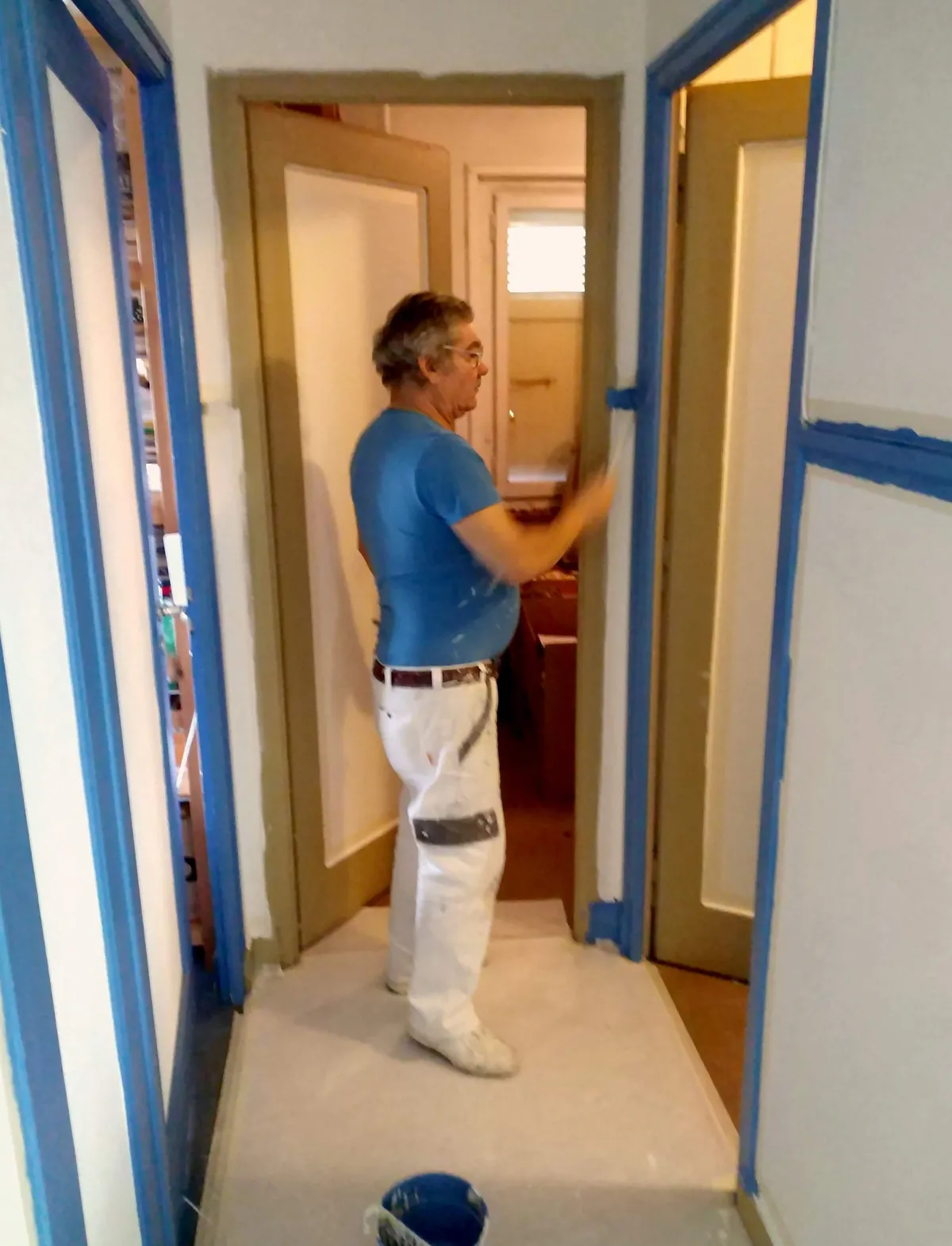 Peintre dans un couloir en train de finir le chambranle bleu d'une porte, une mise en peinture soignée dans les moindres détails.
