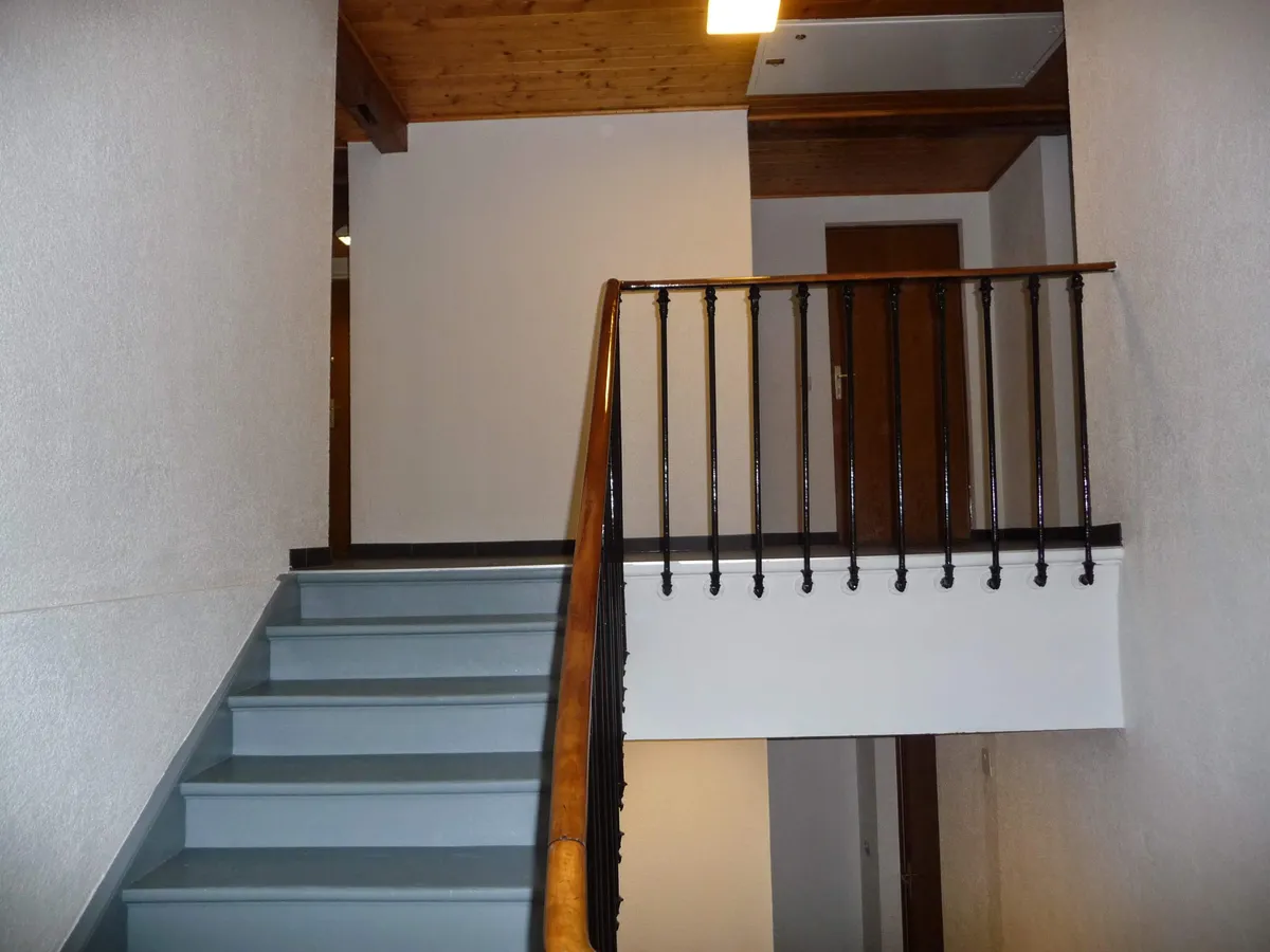 Cage d'escalier aux murs peints, avec un plafond à poutres de bois et une rampe en fer forgé.