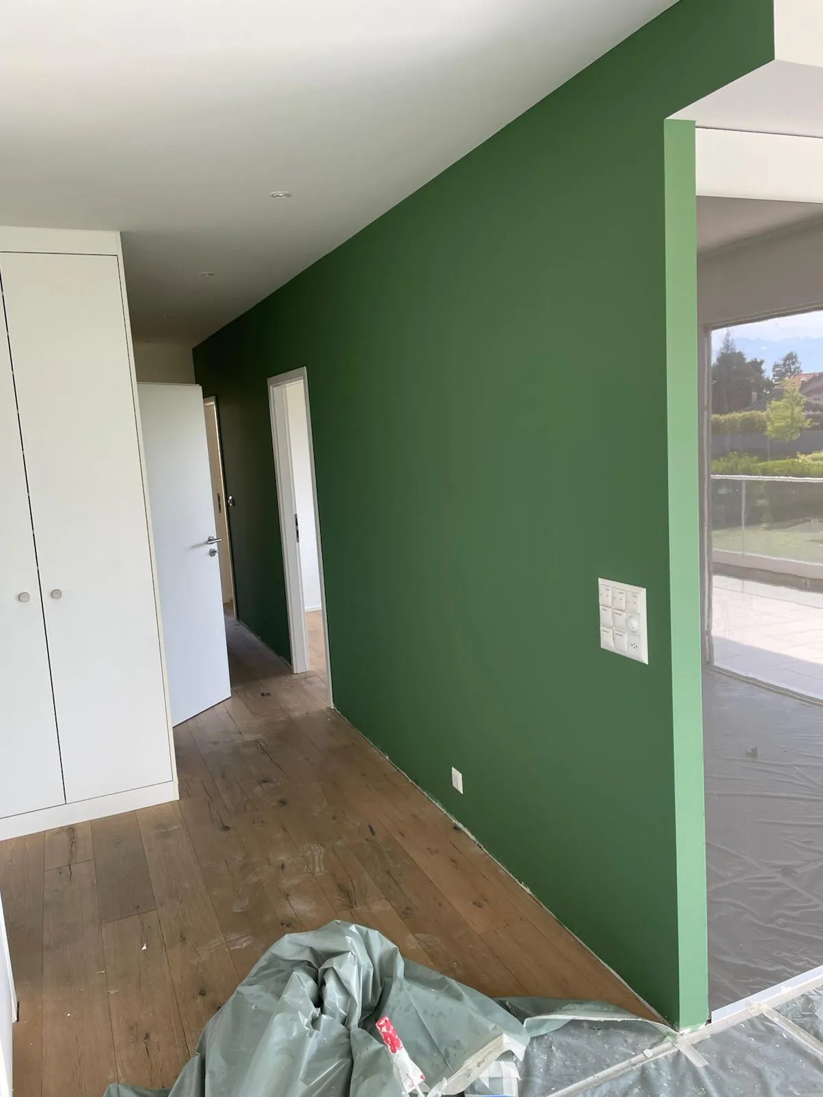 Couloir à mur vert foncé mat, parquet en chêne et portes blanches, chantier de peinture intérieure encore protégé au sol.