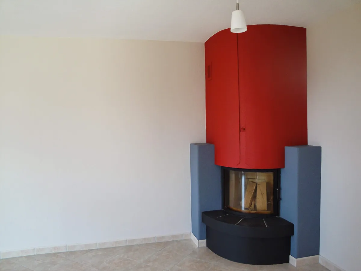 Pièce vide au carrelage clair avec un poêle rouge, murs et plafond repris avant l'emménagement.