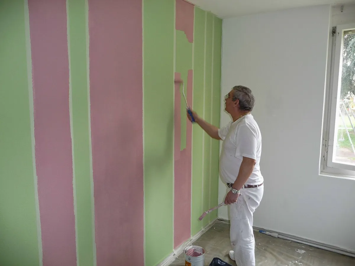 Peintre appliquant des bandes roses entre des bandes vertes sur un mur intérieur.