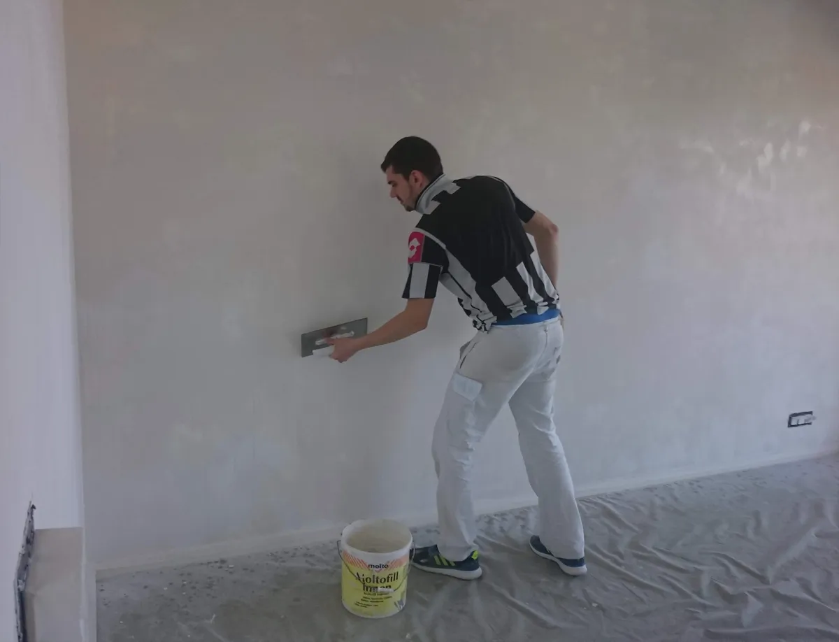 Ouvrier lissant un mur en hauteur à la spatule, avec un seau d'enduit posé au sol.